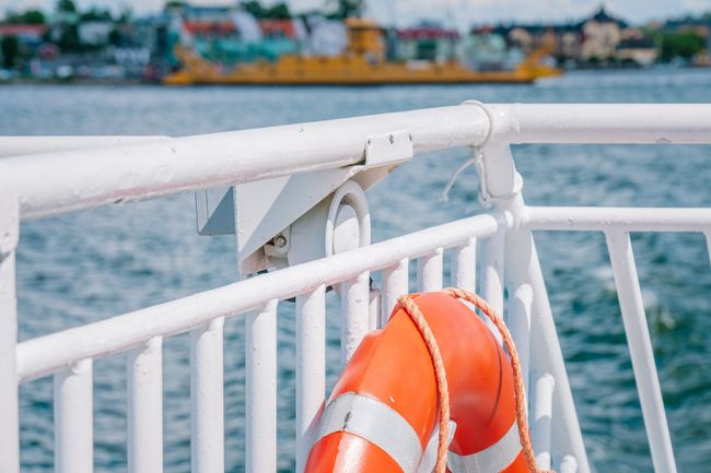 Orange lifebuoy with reflective strips hanging on white railing of ferry boat sailing on blue water with cityscape and yellow ferry boat in the blurry background