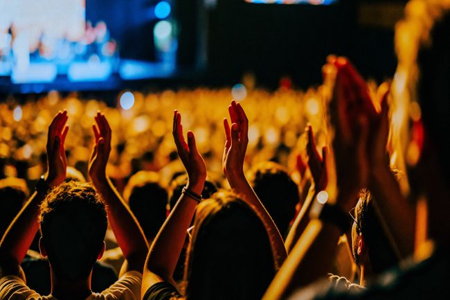 A crowd of people silhouetted against bright stage lights, enthusiastically clapping their hands high above their heads during an exciting live event or music festival.