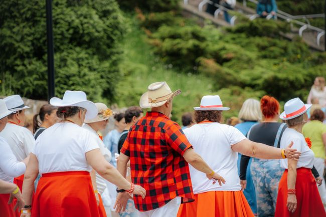 Women dressed in white shirts and red skirts dance together outdoors, wearing hats, with greenery and onlookers visible in the background
