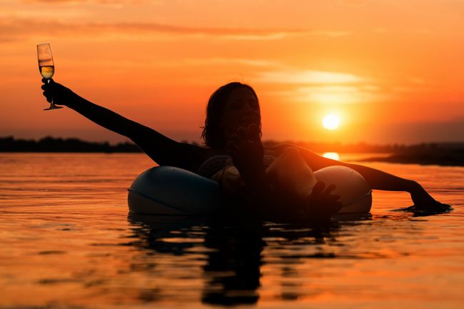 Woman with a glass of champagne lying on inflatable ring in water at sunset or sunrise