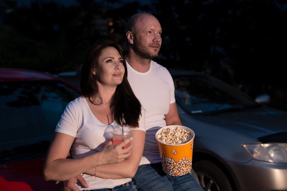 Couple with popcorn standing at the car while watching a movie at drive in cinema. Selective focus. Dating in drive in theater. Entertainment, leisure activities, hobby concept