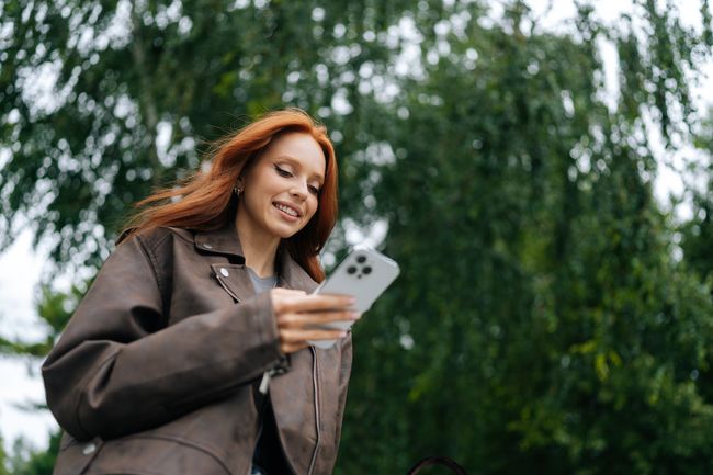 Low-angle view of young woman with long red hair joyfully texting on mobile phone while strolling through city street on summer day. Cheerful redhead female happily messaging on smartphone outdoors.