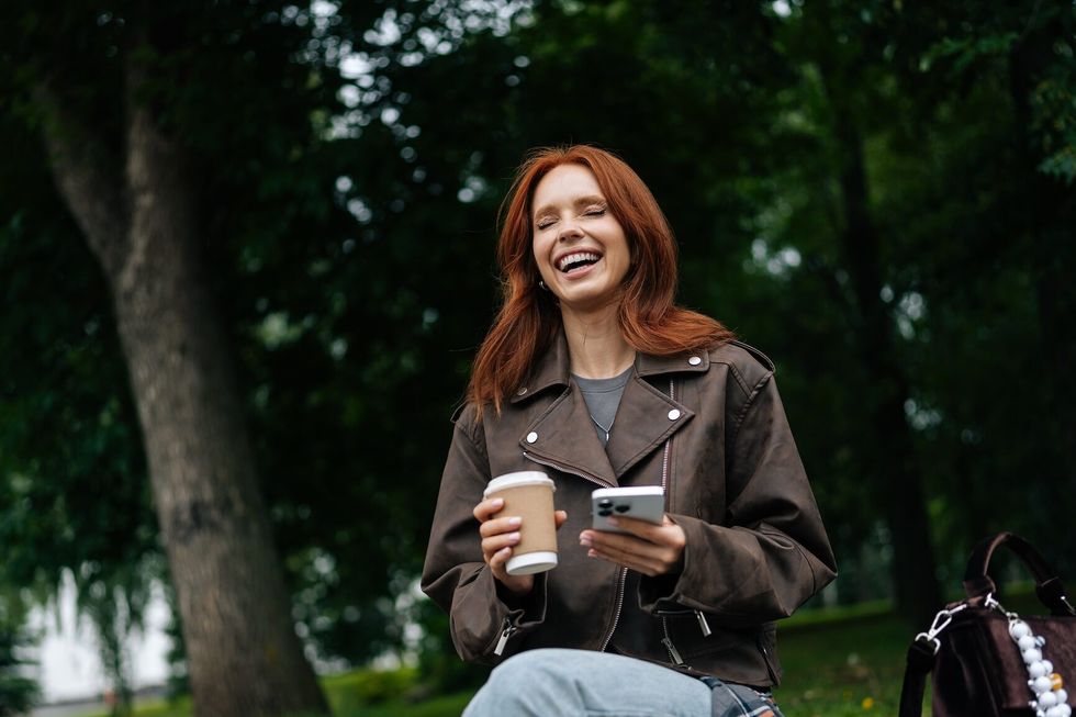 Low-angle view of laughing redhead young woman using smartphone while sitting on bench outdoors in park, enjoying fresh air and greenery on summertime outdoors.