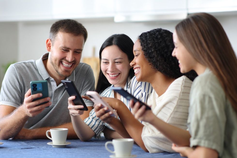 Four happy friends standing in the kitchen checking phones at home