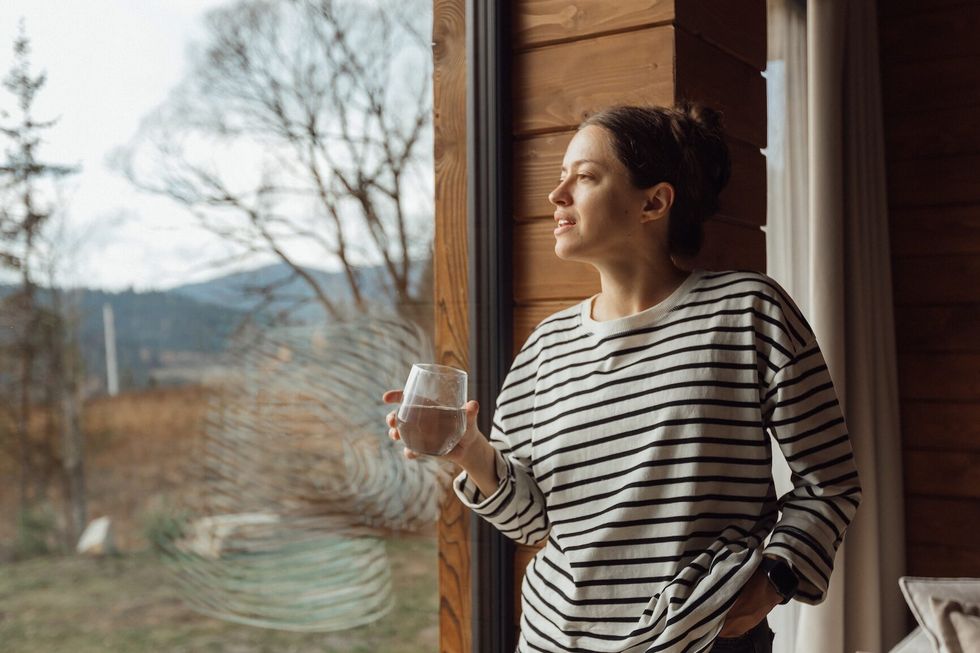 A woman sits comfortably on a gray couch, focusing on her laptop while enjoying a peaceful view of nature through large windows in a wooden cabin living room.