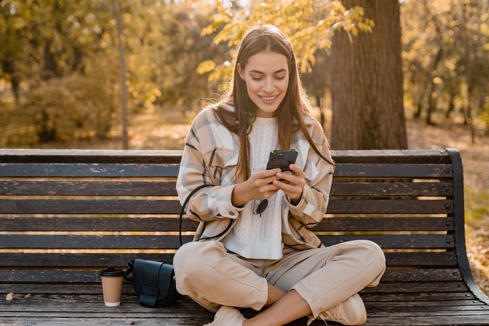candid attractive young smiling woman sitting on bench in autumn park using phone wearing checkered coat, happy mood, fashion style trend