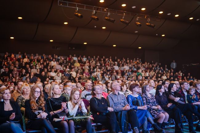audience in a theater with rows of seated people looking towards a stage, attentively watching a performance.