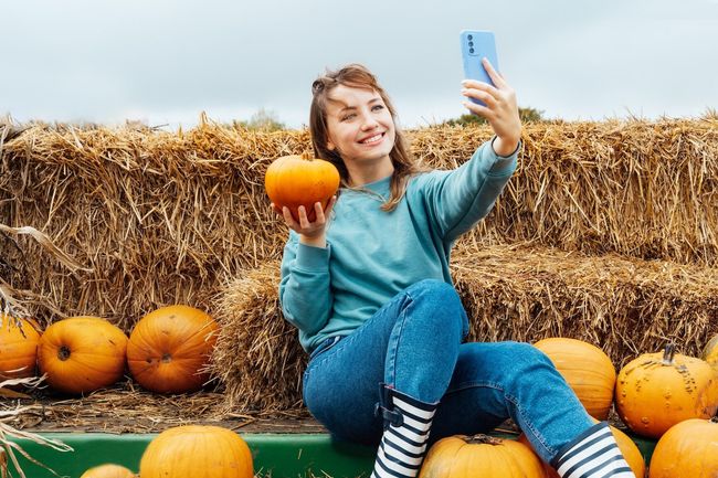 Smiling woman sitting on straw bales and holding pumpkin and making selfie on phone. Selecting Thanksgiving and Halloween holidays decor on agriculture farm. Autumn fall festive mood