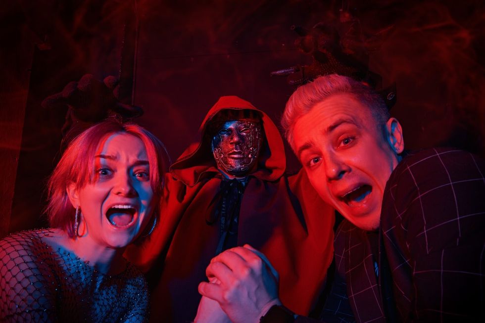 Train of Terror Scary Quest. Two frightened young people are standing in a dark room and a terrible maniac in a black cloak and mask is sneaking behind them. Horror, thriller. Halloween.