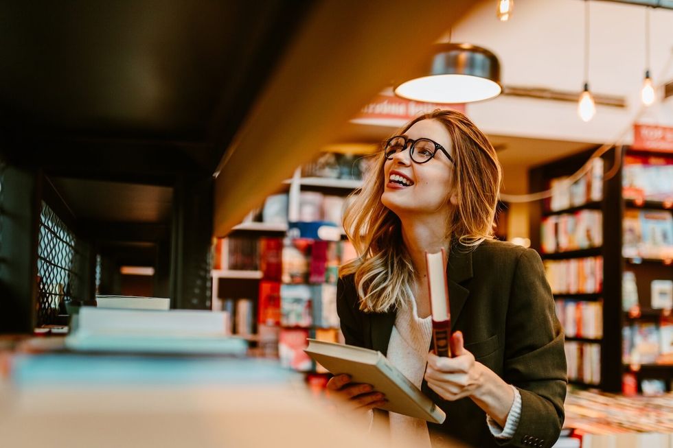 Photo of young woman with glasses returning book to the book shelf. Young woman is looking for book to buy at bookstore.