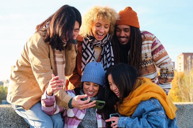 Group of young people laughing and watching something at phone. Smiling multicultural friends looking social media outside the university campus. Students in their free time.
