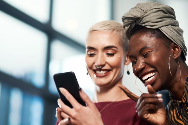 Business, women and pointing with laughing at phone for social media meme, internet joke or funny text message. Creative designer, team and comedy video on smartphone with streaming service in office