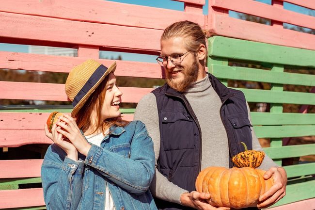 Cheerful young adult man and woman standing near fence, looking to each other and holding ripe, orange pumpkin in hands. People spending free weekend time together, making smiling face