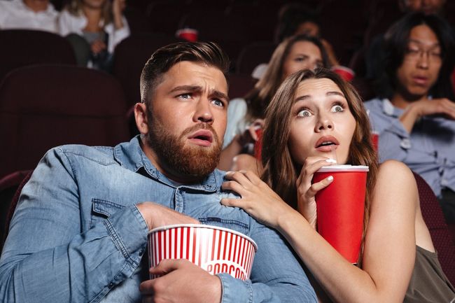 Young scared couple watching a horror movie while sitting at the cinema with popcorn
