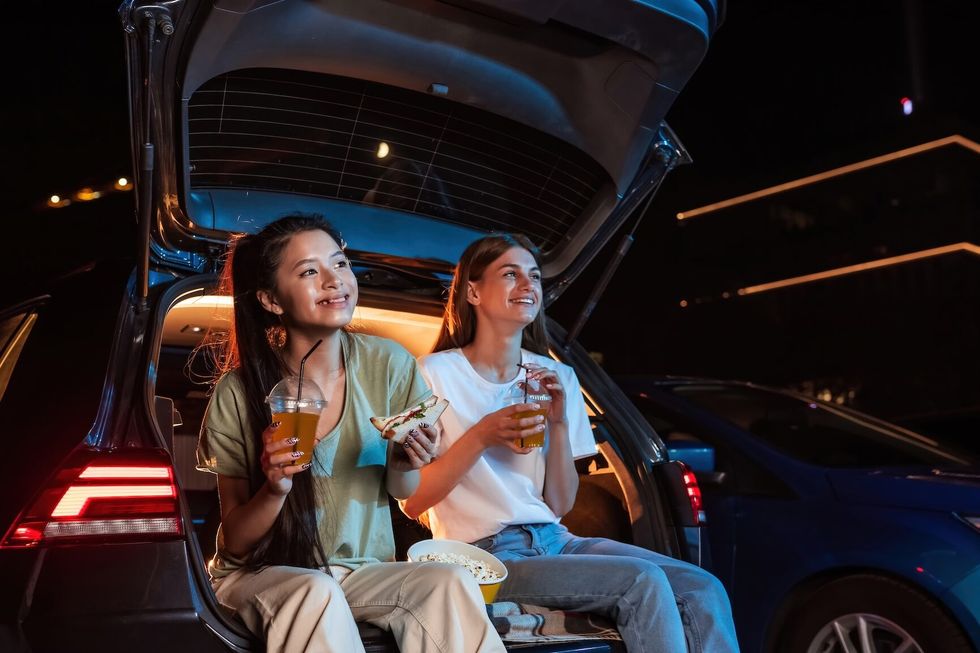 Two happy female best friends smiling while spending time together, having snacks and watching a movie in an outdoor cinema in the evening. Entertainment, friendship concept. Horizontal shot